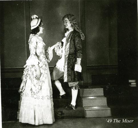 Two actors in 17th-century attire perform a scene from a play. The female actor wears a long, ornate dress and bonnet, while the male actor dons a frock coat and breeches. They are on stage, with the male actor standing on a step. Text reads "'49 The Miser.