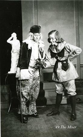 Two actors in vintage costumes perform a scene. One wears a patterned vest and trousers with a hat, while the other, in a tunic and boots, gestures animatedly. A small statue stands in the background. The text "49 The Miser" is in the bottom corner.