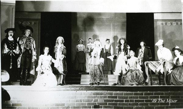 A black and white photo of a theatrical performance with actors in period costumes on stage. The scene features men and women in elaborate outfits, gathered together with one man standing at center stage. The backdrop is simple and the stage is elevated.
