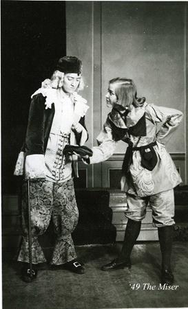 Two actors in period costumes on stage; one in elaborate attire with a cane, the other in simpler clothing. They interact expressively, suggesting a comedic or theatrical scene. A caption reads, "'49 The Miser.