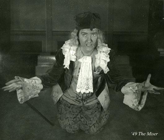 A person dressed in a 17th-century style costume, with a wig, lace cuffs, and a waistcoat, kneels on the floor with arms outstretched. The expression is animated. The image is labeled "'49 The Miser.