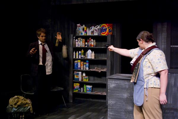 Two actors perform a scene on stage. One holds a gun-like prop, pointing it at the other who has hands raised. Behind them is a shelf with cleaning products and household items. The set is dark with a dramatic ambiance.