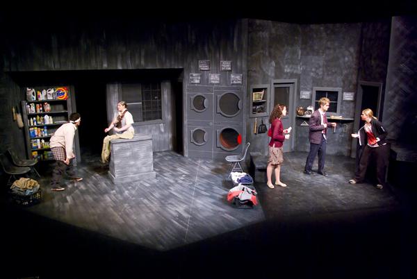 Five actors perform on a stage set resembling a laundromat, with washing machines and detergent shelves. The scene has a monochromatic, gray color scheme. Two actors are sitting, while three are standing, engaging in dialogue.