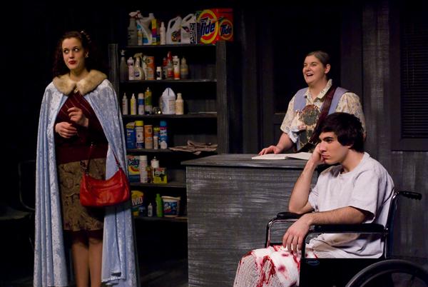 Three people are in a room with shelves holding cleaning supplies. One stands in a fur-trimmed cape holding a bag. Another is smiling behind a counter, while the third person sits in a wheelchair with a bandaged leg, looking pensive.