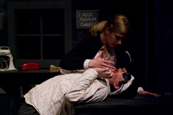 A person in a white jacket and sunglasses lies on a table, while another person, dressed in dark clothing, holds their face gently with one hand. Dim lighting suggests an intense or dramatic scene. A sign reads "1 HOUR MINIMUM CHARGE.