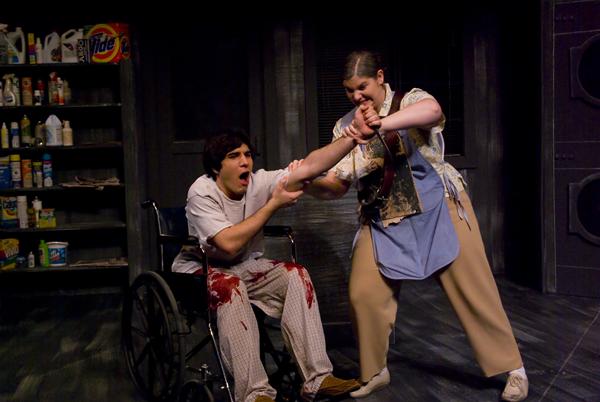 A person in a wheelchair, with blood on their pants, is being restrained by another person gripping their arm. The scene is set indoors with a shelf of household items in the background.