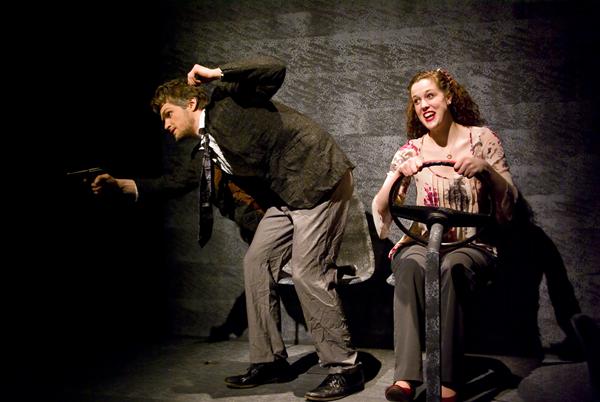 A man and a woman are sitting on chairs mimicking a car scene. The man, wearing a suit, points a toy gun, while the woman, in a patterned blouse, holds a steering wheel and wears a surprised expression. The background is dark and textured.