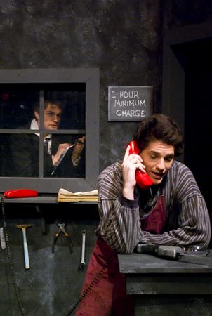 A person leans on a counter, holding a red phone. They wear a striped shirt and apron. In the background, another person peers through a small window. A sign above the window reads "1 hour minimum charge." Tools hang on the wall.