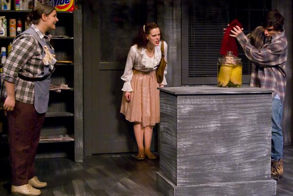 Three actors perform on stage. One actor on the right lifts a red cloth from a large jar on a pedestal. The actor in the center looks surprised, while the actor on the left watches with hands on hips. Shelves with various products are in the background.