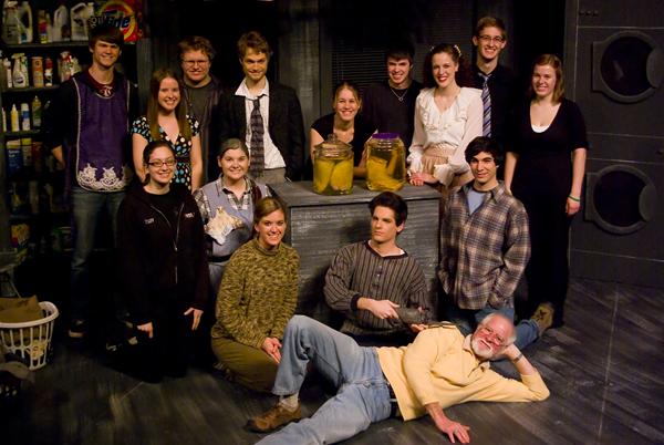 A group of people posing indoors. Some are standing, and others are kneeling or sitting. Two people are sitting around a table with jars on it. Shelves with various items line the wall behind them. Everyone is smiling.
