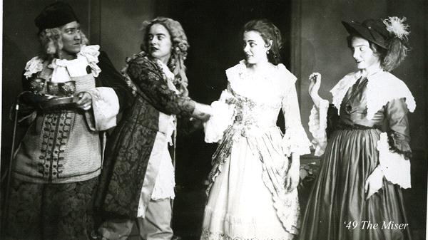 A black and white scene from the play "The Miser" with four actors in 17th-century costumes. One actor offers a tray, another appears to speak, and two women watch. The setting conveys a dramatic moment.