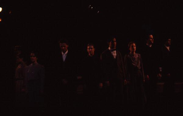 A group of people stands in a dark setting, with minimal lighting highlighting their silhouettes. They appear to be dressed in formal or vintage attire and are arranged in a line facing forward.