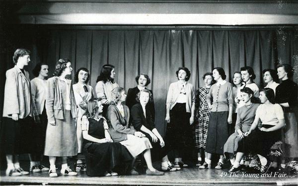 A group of young women are gathered on a stage, some standing and others seated, in front of a curtain. They appear to be engaged in singing or speaking, dressed in mid-20th century attire. The atmosphere suggests a performance or rehearsal.