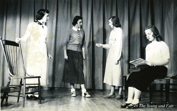 Four women in vintage clothing are on a stage. Three are standing and one is seated reading a book. A backdrop curtain is visible. The setting suggests a theatrical performance or rehearsal.