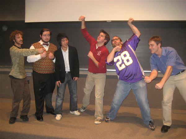 Six people stand on a stage in front of a screen. They are posing playfully, with some making fist gestures and others standing casually. One person is wearing a sports jersey. The group appears to be having fun.