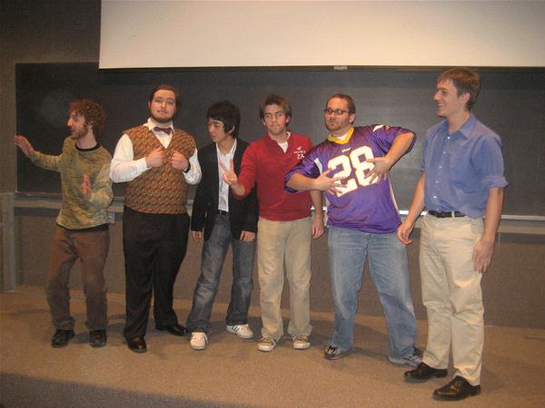 Six men stand on a stage in casual and semi-formal attire, posing playfully. One wears a Viking football jersey, while others sport sweaters and shirts. A chalkboard is visible in the background. They appear to be engaged in a humorous or casual performance.