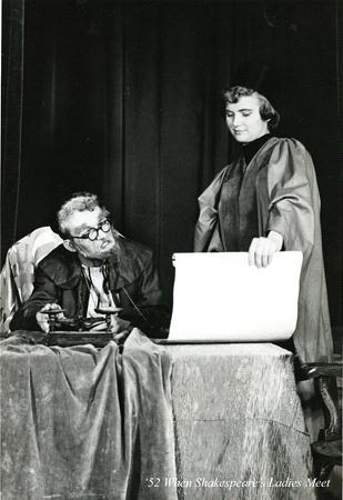 A black and white scene from a play. An older man with glasses sits at a table, looking up, while a woman stands beside him holding a large scroll. The bottom of the image reads "'52 Wigan Shakespeare - Ladies Meet.
