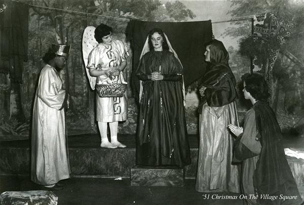 Four people in historical costumes perform a nativity scene with one as an angel and others in robes. They stand on a stage with a woodland backdrop. It's labeled '51 Christmas On The Village Square.