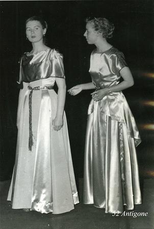 Two individuals in flowing, shiny garments stand against a dark backdrop. One faces forward with a rope belt, while the other turns slightly to look at her. The text "52 Antigone" is visible at the bottom.