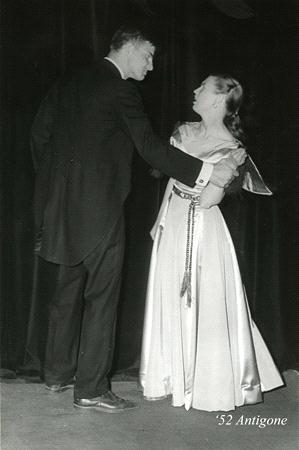 A man and a woman are standing and facing each other on stage, both dressed formally. The woman is wearing a gown and the man is in a suit. The text "'52 Antigone" is in the bottom right corner.