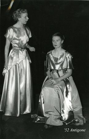Two women in satin dresses perform in a theater production of Antigone, 1952. The standing woman gestures expressively, while the seated woman looks forward with a calm expression. Both are illuminated against a dark backdrop.
