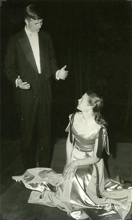 A woman in a long dress is kneeling on the ground, looking up at a standing man in a tuxedo. The scene appears to be theatrical, with both appearing engaged in conversation on a dimly lit stage.