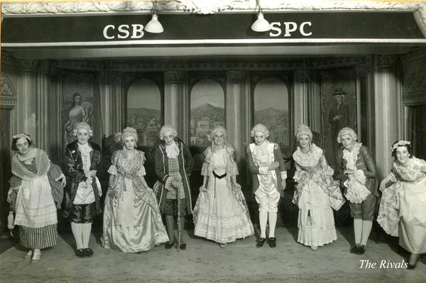 Black and white photo of a group of people in 18th-century period costumes posing on a stage. They are under a banner with the letters "CSB" and "SPC." The backdrop features an ornate architectural setting. The words "The Rivals" are in the corner.