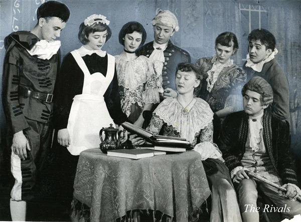 A group of eight people in 18th-century costumes, gathered around a table with books and a teapot. One person is reading from the book. The scene is from "The Rivals.