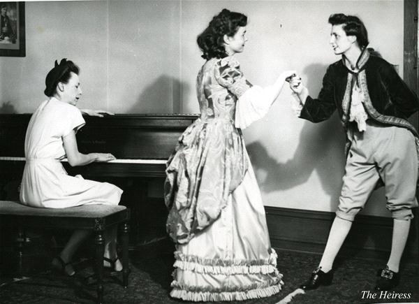 A black and white image of three women in a room. One sits playing the piano, while the other two, dressed in period costumes, are involved in a playful interaction. The woman on the right bows slightly as they hold hands. "The Heiress" is labeled.