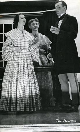 Three people in period costumes are performing a scene on stage. One woman wears a checkered dress, another in a lace dress stands behind, and a man in a suit gestures with his hand. Text reads "The Heiress" in the corner.