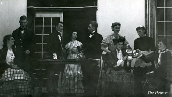 A vintage photo of eight people dressed in period costumes, likely from a theater play. They are gathered around a piano, with two women seated, one holding a parasol. A mix of men and women stand and lean nearby, engaging in conversation.