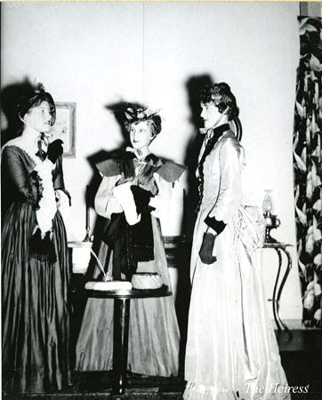 Three women in period costumes from the late 1800s stand in a room with ornate furniture. They wear long dresses, hats, and gloves, engaging in conversation. The room is decorated with a curtain, side table, and lamp. The text "The Heiress" is visible.
