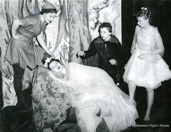 Four actors in theatrical costumes perform a scene from "A Midsummer Night's Dream." One is reclining on a decorated platform, while the others stand around, wearing whimsical attire, with a woodland-themed backdrop.