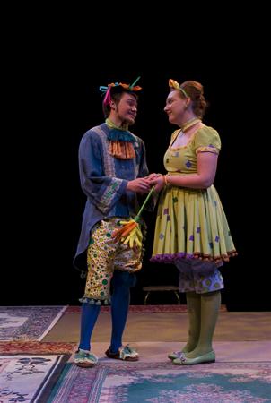 Two actors in colorful costumes hold hands on stage. The man wears a blue tunic and patterned pants, while the woman is in a green polka-dot dress. Both have decorative headgear. They stand on a carpeted floor against a black background.