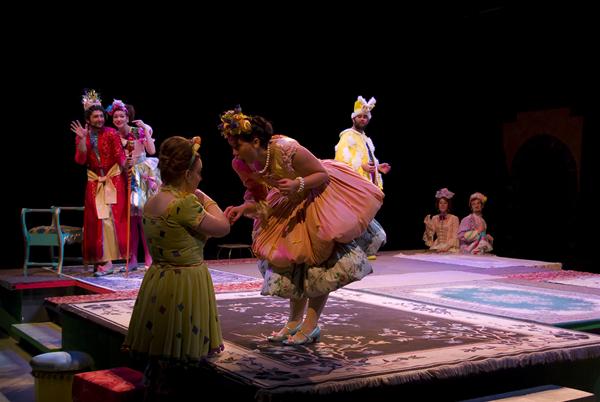 A theatrical performance with actors in colorful, elaborate costumes. One actor, in a pink dress, leans forward to kiss another's hand. Others in the background wear crowns and diverse attire, all on a carpeted stage with dim lighting.
