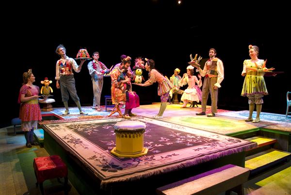 A colorful theater stage with eight actors in whimsical costumes perform a play. The stage is decorated with a patterned rug and various props. Some actors hold trays or wear playful hats, creating a lively, theatrical atmosphere.
