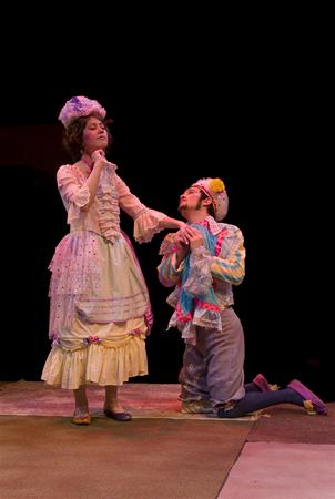 Two people in period costumes on stage. One stands, wearing a white, ruffled dress and hat, while the other kneels, holding the first person's hand, dressed in a colorful outfit with a hat. Dimly lit theatrical setting.