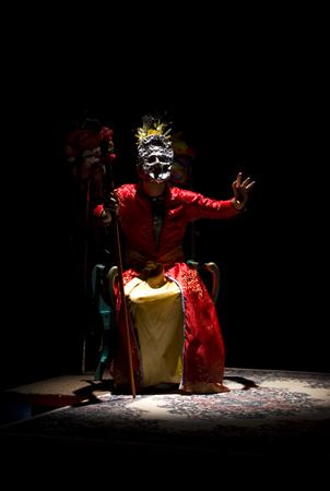 Person in a red costume and elaborate mask sits on a stage under a spotlight, holding a staff. The setting is dark, and a patterned rug is visible beneath them. They have one hand raised, adding a dramatic effect.