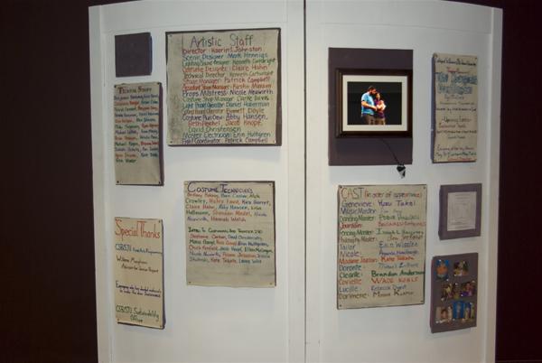 A display board featuring handwritten lists for artistic staff, special thanks, and cast members. It includes a framed photo at the top right and several small cards and images on the lower right section of the board.