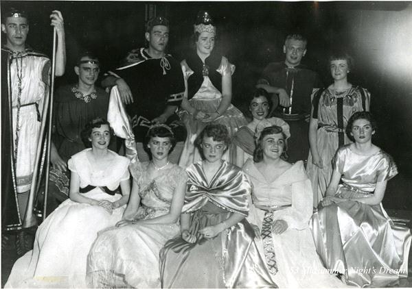 A group of men and women in medieval costumes pose for a photo. Some are seated, others standing, wearing gowns, robes, and crowns. The setting appears to be a theatrical production, possibly of a Shakespearean play.