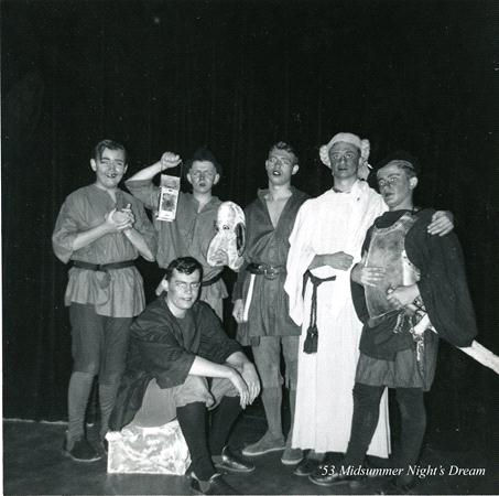 A group of six men in costume pose for a photo on stage. They are dressed in medieval-style outfits. One man sits on a box, while the others stand around him, holding masks and props. The text on the image reads "'53 Midsummer Night's Dream.