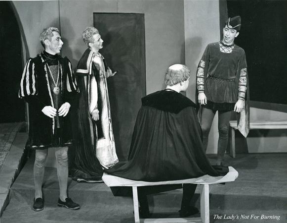 Black and white photo of four actors on stage in period costumes. One actor sits on a bench with his back to the camera, while the others stand, seemingly in mid-conversation. A caption reads "The Lady's Not for Burning.