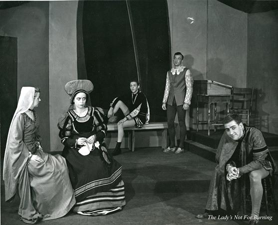 A black and white photo of five actors in period costumes performing in a stage play, with three sitting and two standing. The set includes a bench and chairs. The caption reads "The Lady's Not For Burning.
