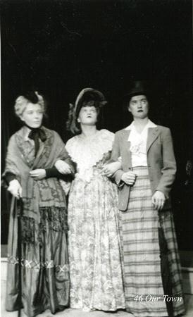 Three people dressed in vintage clothing stand together on stage. The person on the left wears a shawl, the middle person wears a patterned dress and large hat, and the person on the right wears a blazer, tie, and plaid skirt. The image is in black and white.
