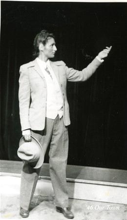A person wearing a suit and vest stands on stage against a dark curtain, holding a hat in one hand and gesturing with the other. The setting is theatrical, suggesting a scene from a play.