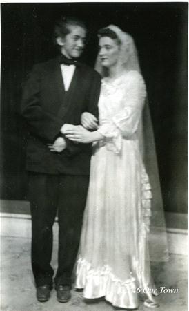 A black and white photo of two people in formal attire. One wears a dark suit with a bow tie, and the other is in a long white gown with a veil. They are standing close, holding arms, and smiling. "46 Our Town" is written in the corner.