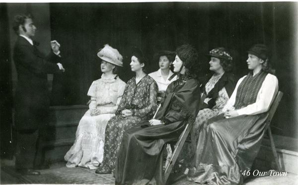 A black and white photo of six women in period costumes seated in a row with one man standing to the left, appearing to speak or direct them. The women wear long dresses and large hats. The image is labeled "'46 Our Town.