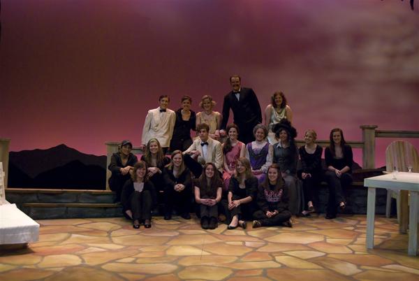 A group of people, some seated and some standing, pose together on a stage with a stylized sunset backdrop. A white table and chair are visible on the right. The group appears relaxed and smiling.