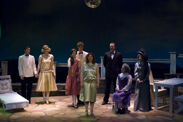 A group of people in formal attire stand on a stage set designed to look like an outdoor patio at night, with a moon in the background. The scene includes tables and chairs, with a woman in the center wearing a light-colored dress.