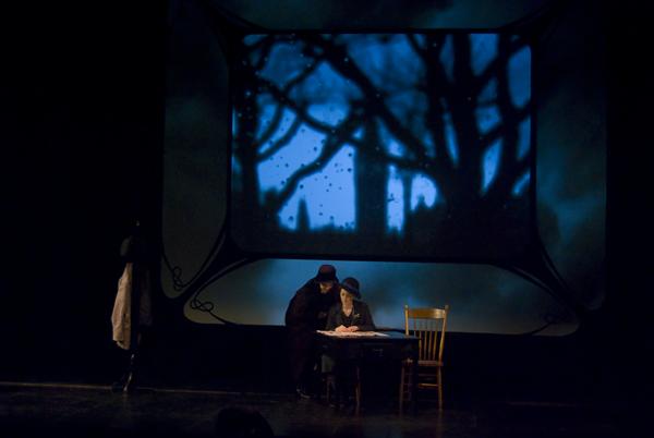 Two figures are on a dimly lit stage. One is sitting at a table with an open book, while the other leans over. In the background, a screen displays a dark, tree-filled scene with a blue hue. A wooden chair is visible beside the table.